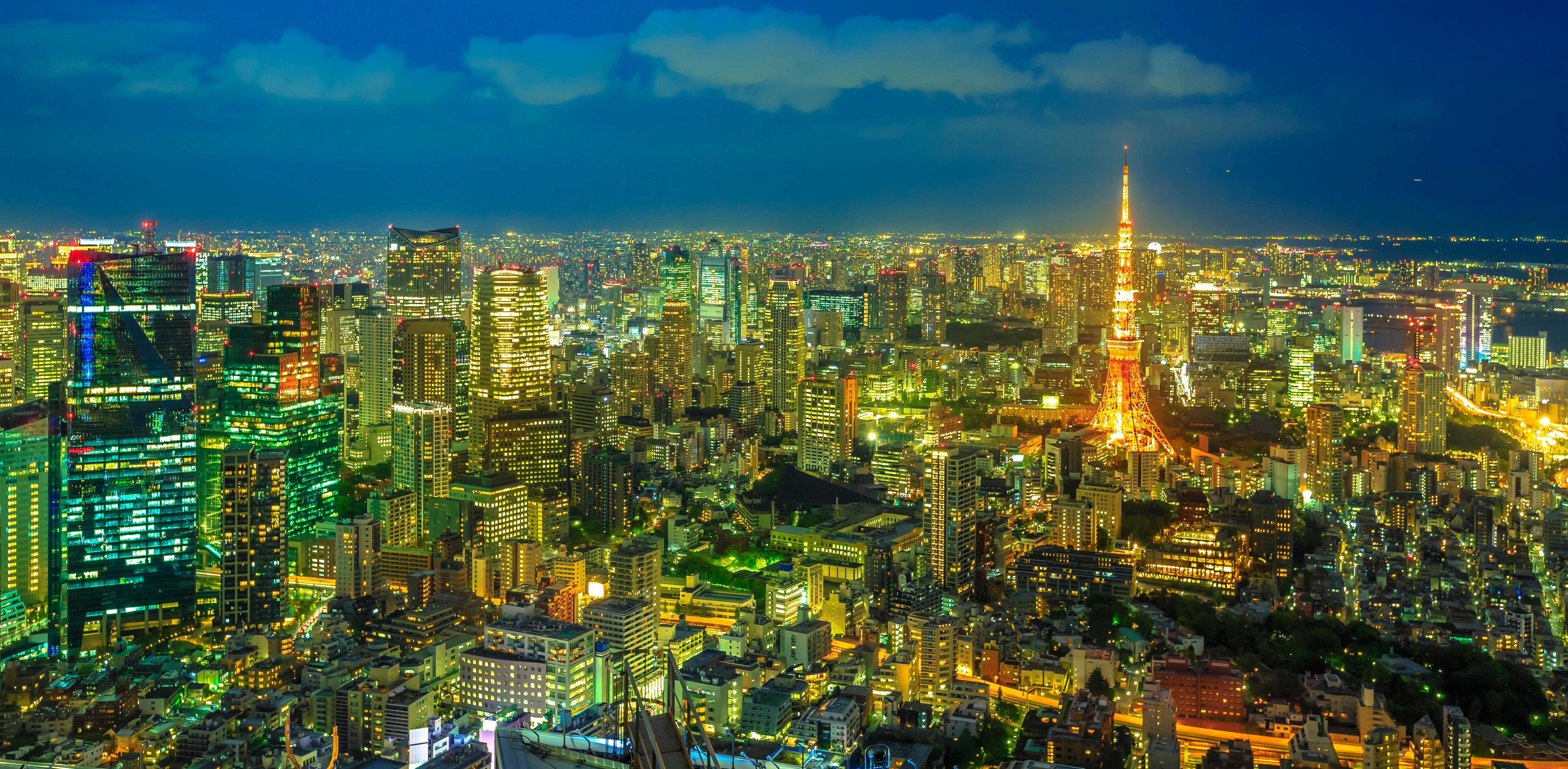 A vibrant cityscape of Tokyo at night with illuminated buildings and Tokyo Tower glowing in orange lights, standing tall among the city lights under a deep blue sky.