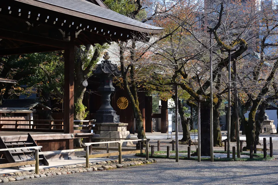 A peaceful view of a traditional wooden Japanese shrine with a courtyard covered in gravel. Leafless trees with sparse branches stand near the shrine, and a stone lantern is visible in the background. The scene is calm and serene.