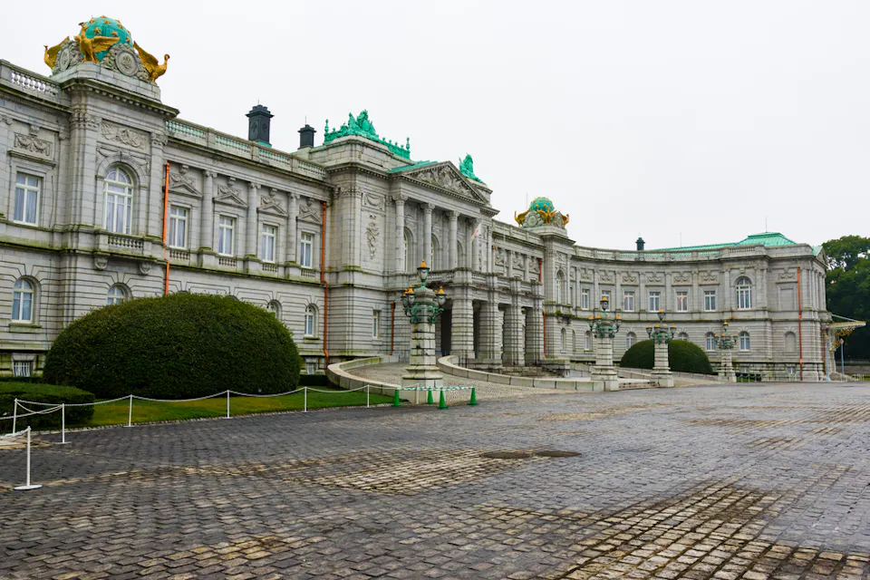 A large, ornate neo-baroque palace with rugged stone walls and detailed sculptures on the roofline. The entrance features grand columns and steps leading to the door. The foreground has a spacious cobblestone courtyard bordered by trimmed hedges.