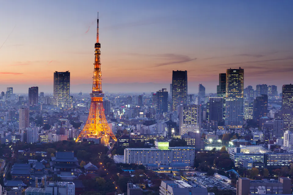 Tokyo skyline at dusk featuring Tokyo Tower illuminated in warm lights. Skyscrapers and buildings surround the tower, while the sky transitions from orange to blue. The cityscape showcases a blend of modern and traditional architecture.