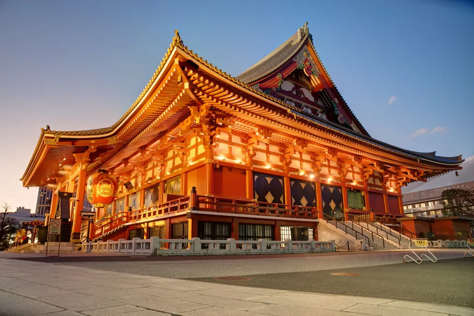 A beautifully illuminated red and gold traditional Japanese temple building with intricate detailing stands majestically against a partly cloudy sky at dusk. The area around the temple is empty, with no people in sight, and the ground is paved.