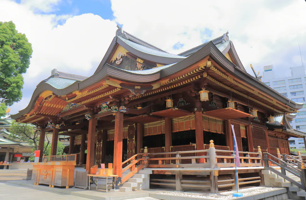 A traditional Japanese Shinto shrine featuring ornate wooden architecture, curved rooflines, and gold accents. The shrine is surrounded by a serene outdoor area with trees and a few modern buildings visible in the background. Bright daylight illuminates the scene.