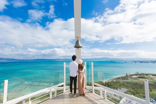 A couple stands on a wooden deck overlooking a bright blue sea, facing a large bell suspended from a white structure, with a partly cloudy sky and distant islands in the background.