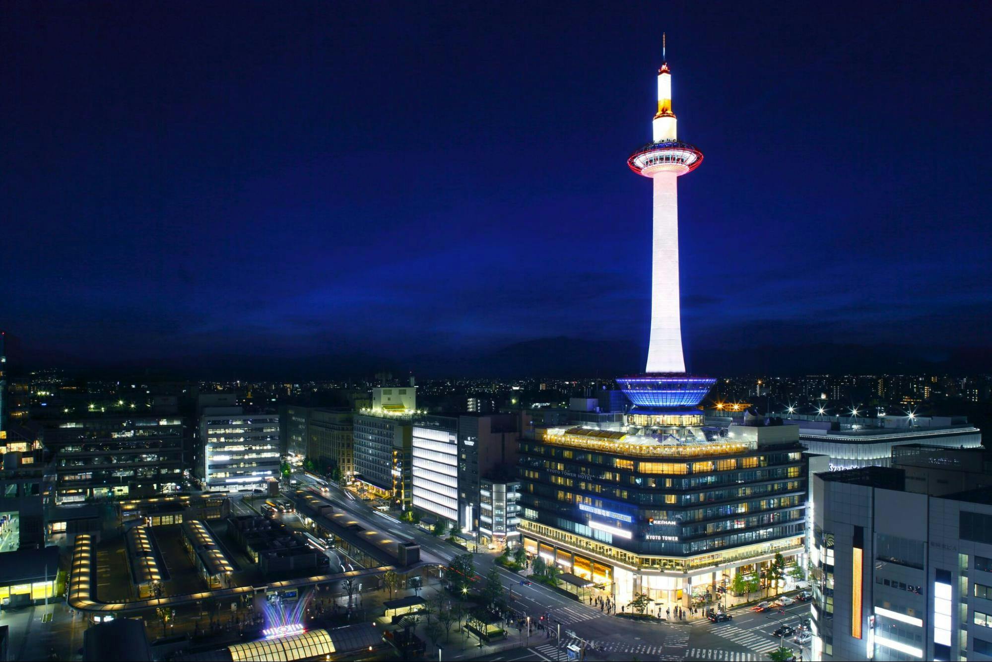 Kyoto Tower illuminated at night, glowing white and orange, stands tall above surrounding city buildings under a deep blue sky in Kyoto, Japan.