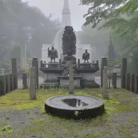 Temple Hidden in Tokyo A misty scene featuring a serene outdoor area in a Japanese temple or shrine. Central monuments include sculptures of figures and a circular stone basin with water. Moss-covered ground and towering trees enclose the space, with a white pagoda in the background.