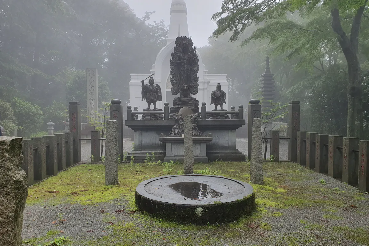 Temple Hidden in Tokyo A misty scene featuring a serene outdoor area in a Japanese temple or shrine. Central monuments include sculptures of figures and a circular stone basin with water. Moss-covered ground and towering trees enclose the space, with a white pagoda in the background.