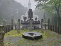 A misty scene featuring a serene outdoor area in a Japanese temple or shrine. Central monuments include sculptures of figures and a circular stone basin with water. Moss-covered ground and towering trees enclose the space, with a white pagoda in the background.