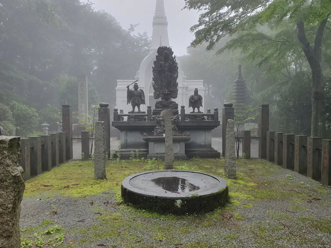 A misty scene featuring a serene outdoor area in a Japanese temple or shrine. Central monuments include sculptures of figures and a circular stone basin with water. Moss-covered ground and towering trees enclose the space, with a white pagoda in the background.
