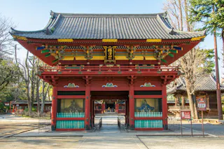 A traditional Japanese temple gate with elaborate red wooden beams, green railings, and ornate decorative accents, standing under a tiled roof on a sunny day. Trees and temple grounds are visible in the background.