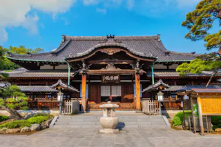 Traditional Japanese temple with a curved tiled roof, wooden exterior, and intricate architectural details, surrounded by greenery and a stone lantern in front under a bright blue sky.
