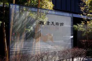A glass sign for the Nezu Museum stands outside, surrounded by bamboo and shrubs, reflecting nearby trees and the street. The sign has Japanese and English text with a modern graphic design.