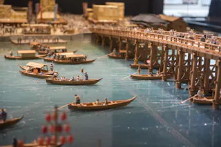 A detailed miniature diorama shows wooden boats with tiny figures rowing on a river, and a busy wooden bridge filled with more miniature people above, recreating a lively historical scene.