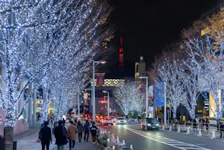 A city street at night lined with trees covered in bright white holiday lights, with people walking on the sidewalks and cars driving on the road. Tall buildings and colorful signs are visible in the background.