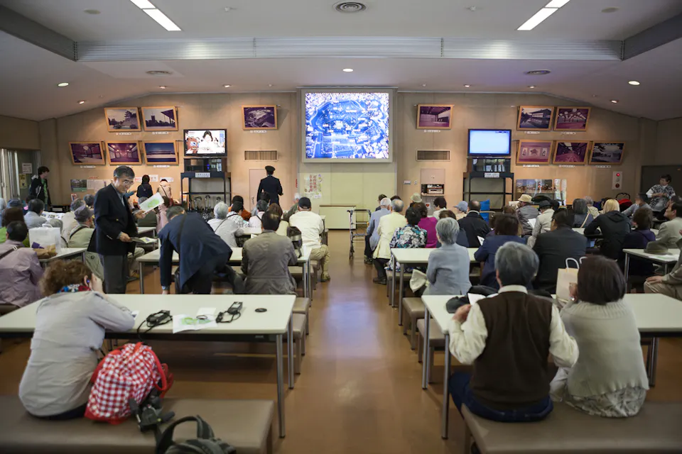 People sitting and standing in a communal room, facing a large screen displaying a cityscape. The room has tables, chairs, and wall displays with various images. The atmosphere appears to be that of a group meeting or presentation.