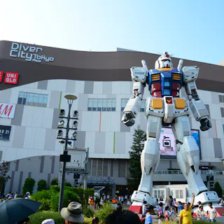 DiverCity Tokyo Plaza in Odaiba A large crowd gathers around a life-size Gundam robot statue in front of the DiverCity Tokyo Plaza shopping mall on a sunny day. The mall’s exterior features various store signs.
