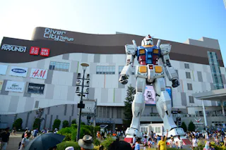 A large crowd gathers around a life-size Gundam robot statue in front of the DiverCity Tokyo Plaza shopping mall on a sunny day. The mall’s exterior features various store signs.
