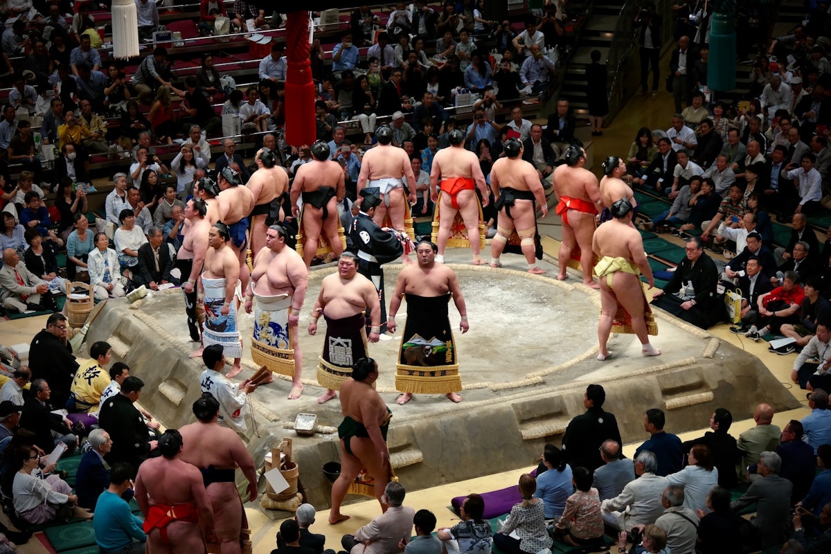 Sumo Wrestlers A group of sumo wrestlers in traditional attire stand and bow in a circular ring surrounded by a large crowd of seated spectators in an indoor arena. The wrestlers are in various positions, with some facing each other and others facing the audience.