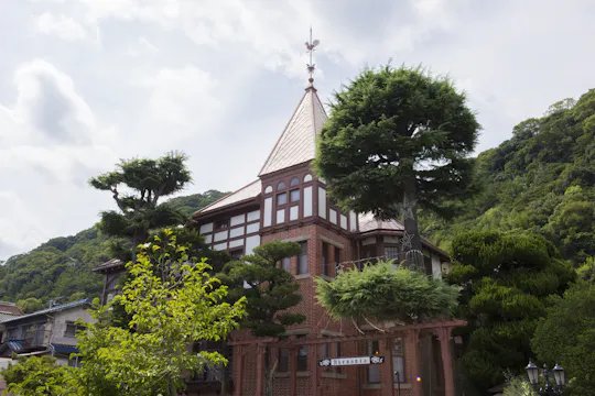 A historic brick building with a steep triangular roof and decorative spire, surrounded by neatly trimmed trees and greenery under a partly cloudy sky.