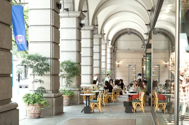 People sit at outdoor tables under stone arches of a building, enjoying food and drinks. Some greenery in large pots decorates the walkway. A server stands near the tables and a blue flag hangs on the left.