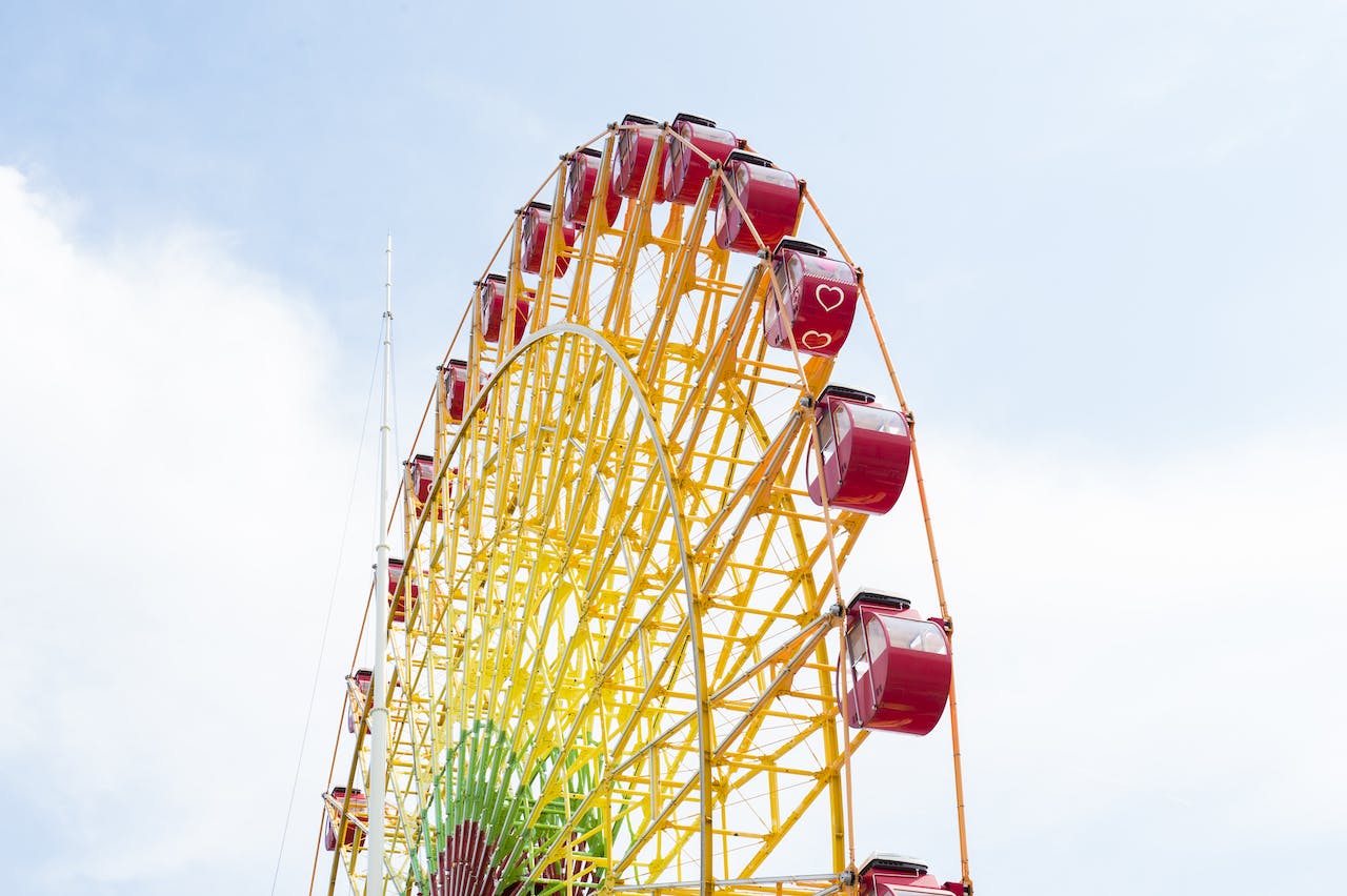 A large yellow Ferris wheel with red gondolas, some decorated with white heart shapes, stands against a bright blue sky with scattered clouds.