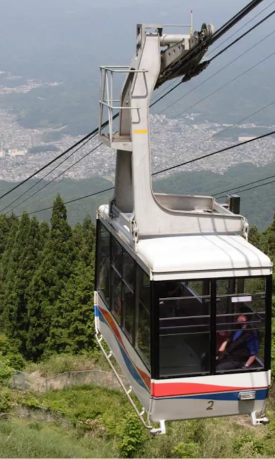 Eizan Cable Car A cable car ascends a green, tree-covered mountain, suspended by cables, with a city visible in the hazy distance below.