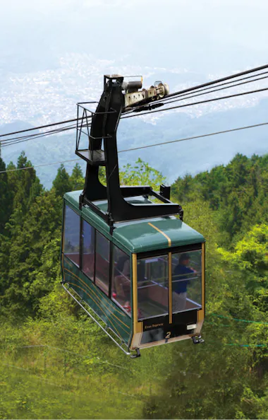 Eizan Cable Car A green cable car travels over a lush, green mountainside surrounded by dense trees, with distant cityscape and hills visible through the haze in the background.
