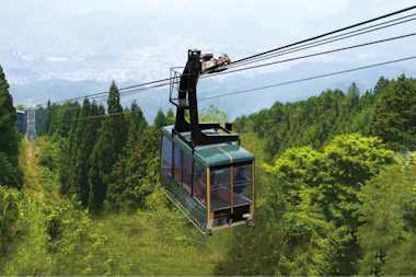 A green cable car travels over a lush, green mountainside surrounded by dense trees, with distant cityscape and hills visible through the haze in the background.