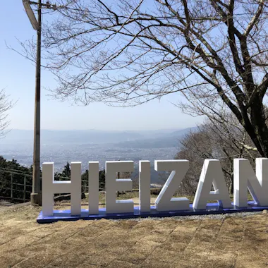 Eizan Cable Car Large white letters spelling "HIEIZAN" stand outdoors on a hillside, with leafless trees and a hazy cityscape visible in the background under a clear sky.