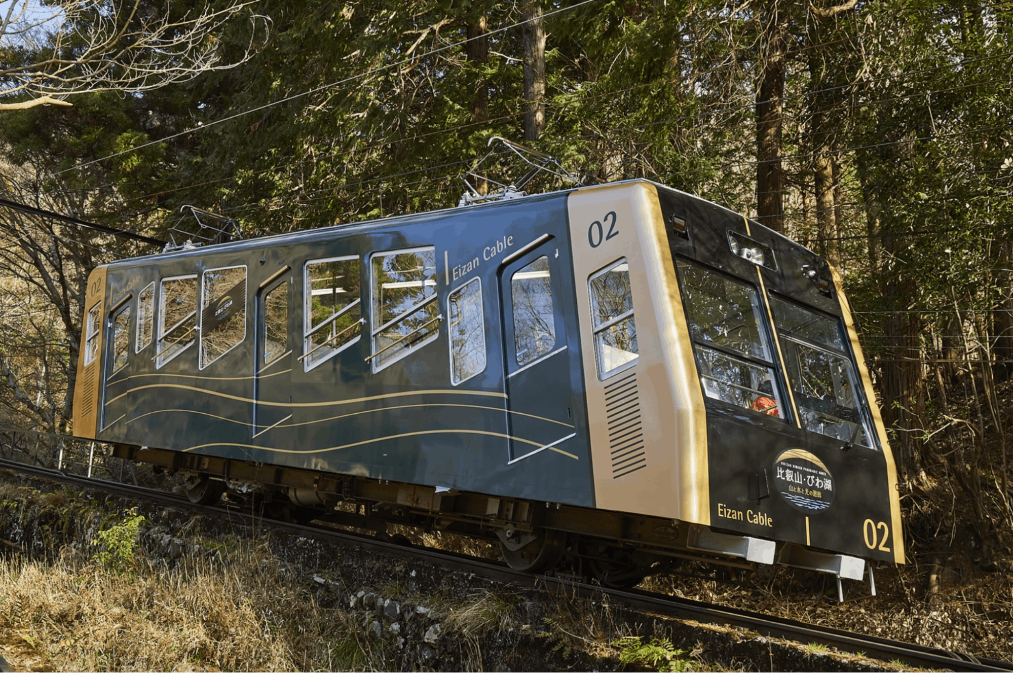 A blue and gold cable car labeled "Eizan Cable 02" travels uphill on a steep track through a forested area, surrounded by trees and greenery.