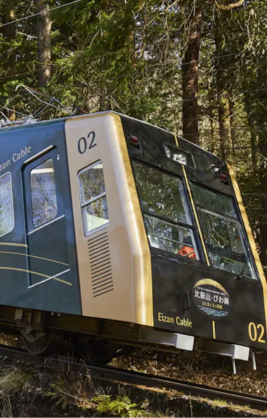 Eizan Cable Car A blue and gold cable car labeled "Eizan Cable 02" travels uphill on a steep track through a forested area, surrounded by trees and greenery.