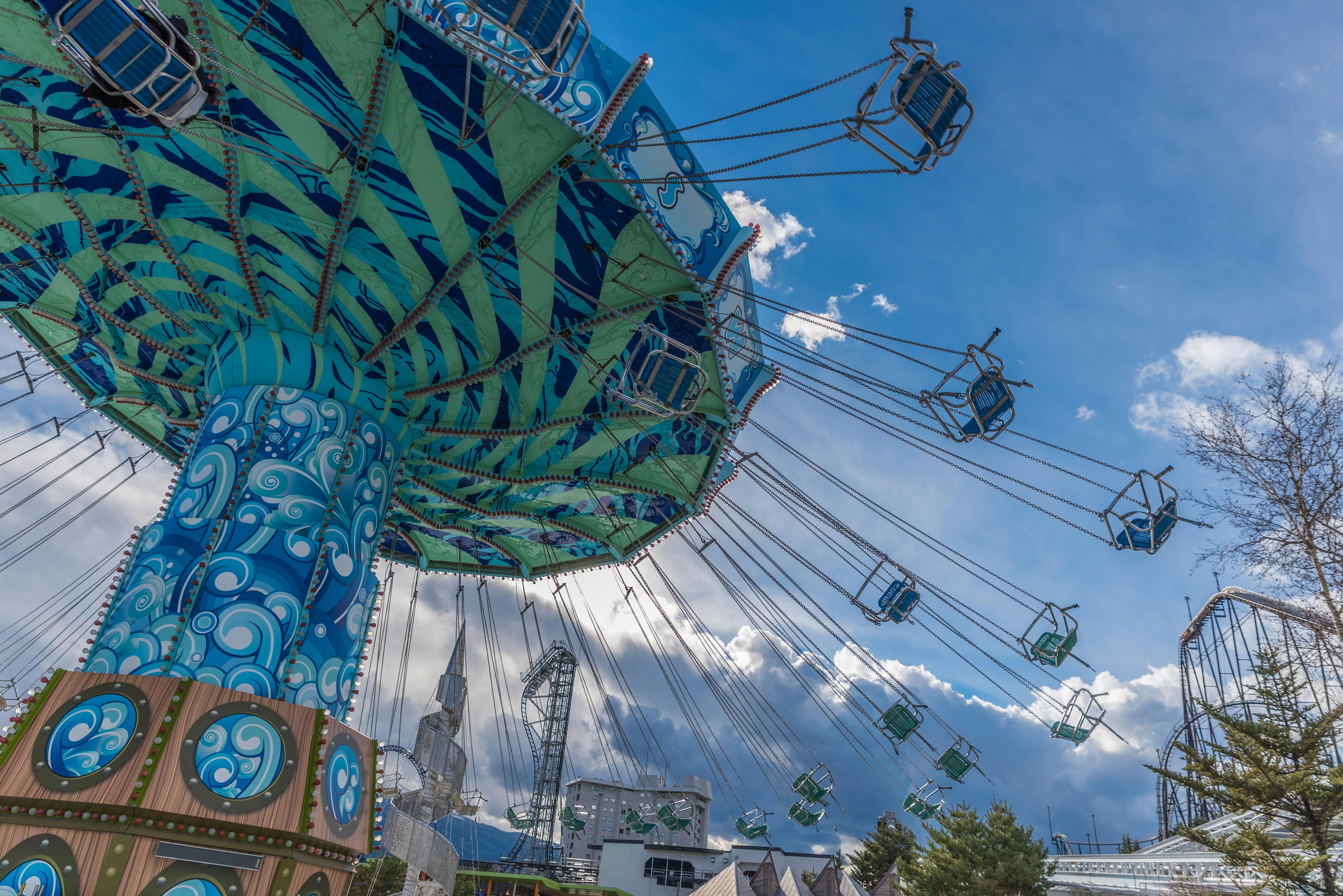 Colorful swing ride at an amusement park, empty blue chairs hanging from chains, set against a bright blue sky with clouds. Some trees and roller coaster tracks are visible in the background.