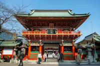A group of people stands in front of a traditional Japanese shrine with a green-tiled roof, red columns, ornate decorations, and a clear blue sky in the background.