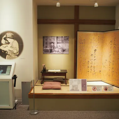 A museum display features a traditional Japanese tatami mat area with a low desk and cushion, large calligraphy panel, old photographs, and informational signs. Warm lighting highlights the exhibit’s cultural artifacts.