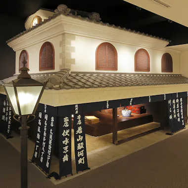 A traditional Japanese building exhibit with a tiled roof, wooden shutters, and fabric banners with white kanji. Inside, wooden shelves display various items. A vintage street lamp stands near the entrance.