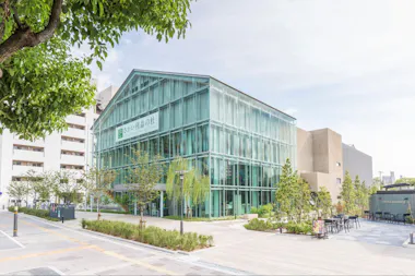 A modern, two-story building with glass walls stands on a clean, tree-lined street. The building is surrounded by greenery, and there are outdoor tables and chairs nearby under a partly cloudy sky.