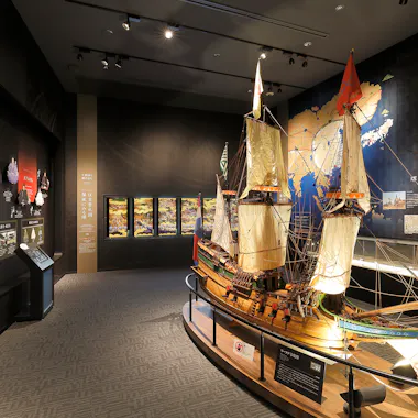 A museum exhibit room featuring a large detailed model of a historic sailing ship, information panels, illuminated displays, and a world map mural on the wall.