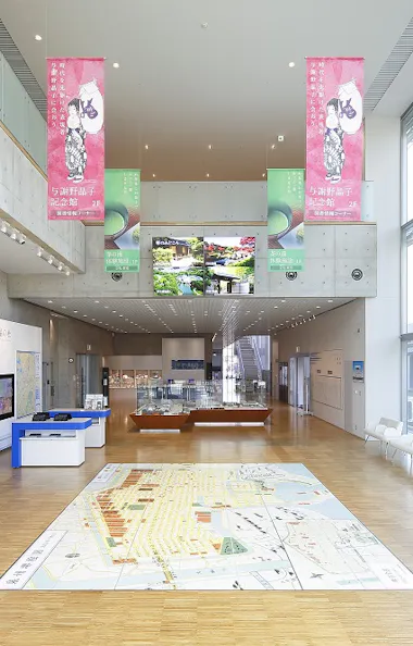 Sakai Plaza of Rikyu and Akiko Spacious, modern museum interior with large windows, wall maps, interactive screens, and a city map on the floor. Pink banners with illustrations hang from the ceiling, and digital displays are visible above the main entrance.