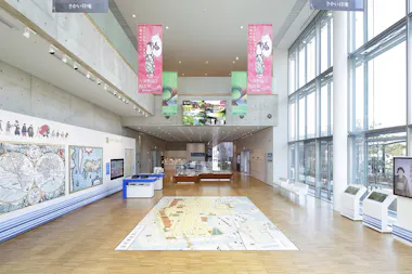 Spacious, modern museum interior with large windows, wall maps, interactive screens, and a city map on the floor. Pink banners with illustrations hang from the ceiling, and digital displays are visible above the main entrance.