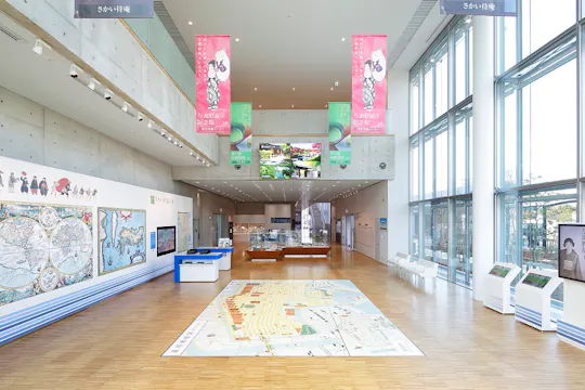 Spacious, modern museum interior with large windows, wall maps, interactive screens, and a city map on the floor. Pink banners with illustrations hang from the ceiling, and digital displays are visible above the main entrance.