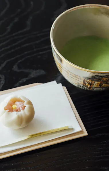 Matcha and Wagashi A traditional Japanese sweet and a small utensil are placed on a white napkin atop a wooden tray next to a bowl of green matcha tea on a dark, textured table.