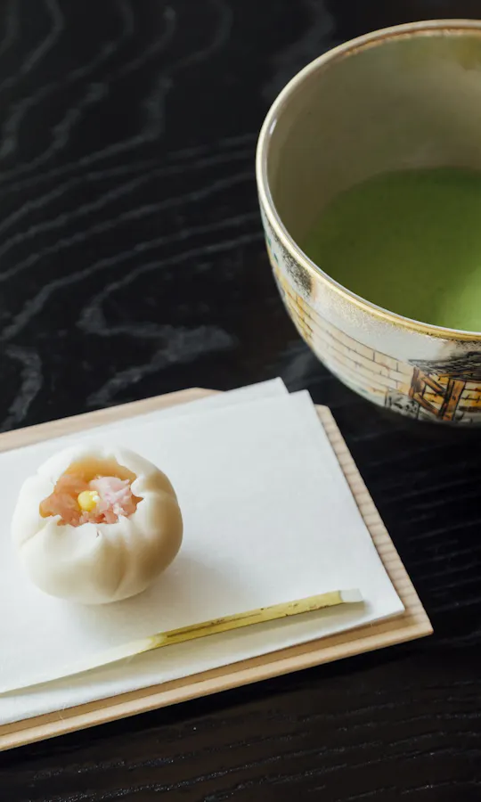 Matcha and Wagashi A traditional Japanese sweet and a small utensil are placed on a white napkin atop a wooden tray next to a bowl of green matcha tea on a dark, textured table.