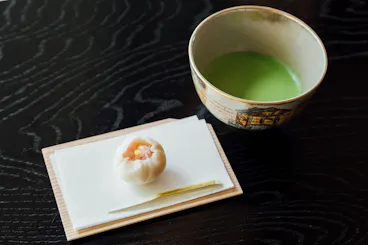 A traditional Japanese sweet and a small utensil are placed on a white napkin atop a wooden tray next to a bowl of green matcha tea on a dark, textured table.