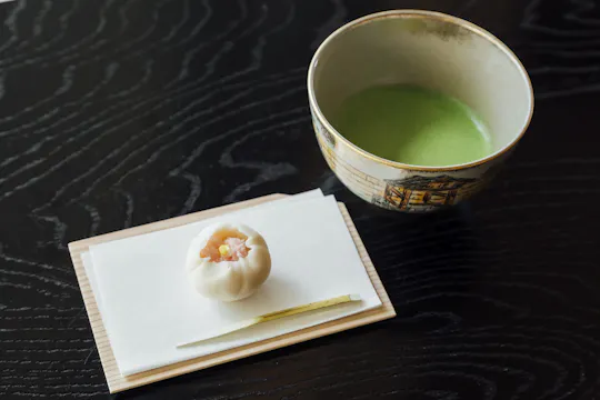 A traditional Japanese sweet and a small utensil are placed on a white napkin atop a wooden tray next to a bowl of green matcha tea on a dark, textured table.