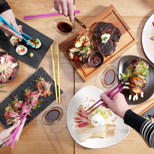 Japanese Food Top view of a dining table with various Asian fusion dishes, including sushi, grilled meat, and salads. Several hands using colorful chopsticks are reaching for the food, showcasing a shared meal experience.