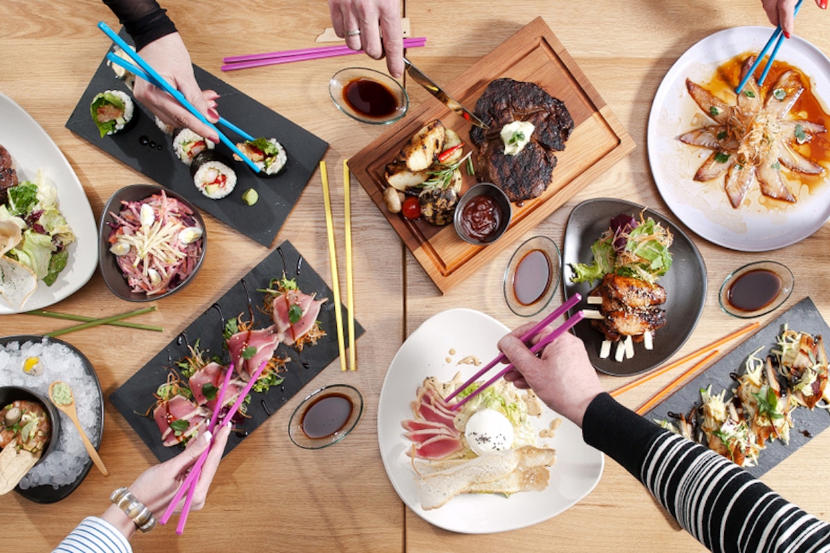Japanese Food Aerial view of a dining table with various dishes, including sushi, grilled meats, salads, and desserts. Multiple hands using chopsticks reach for the food, creating a lively and colorful scene.