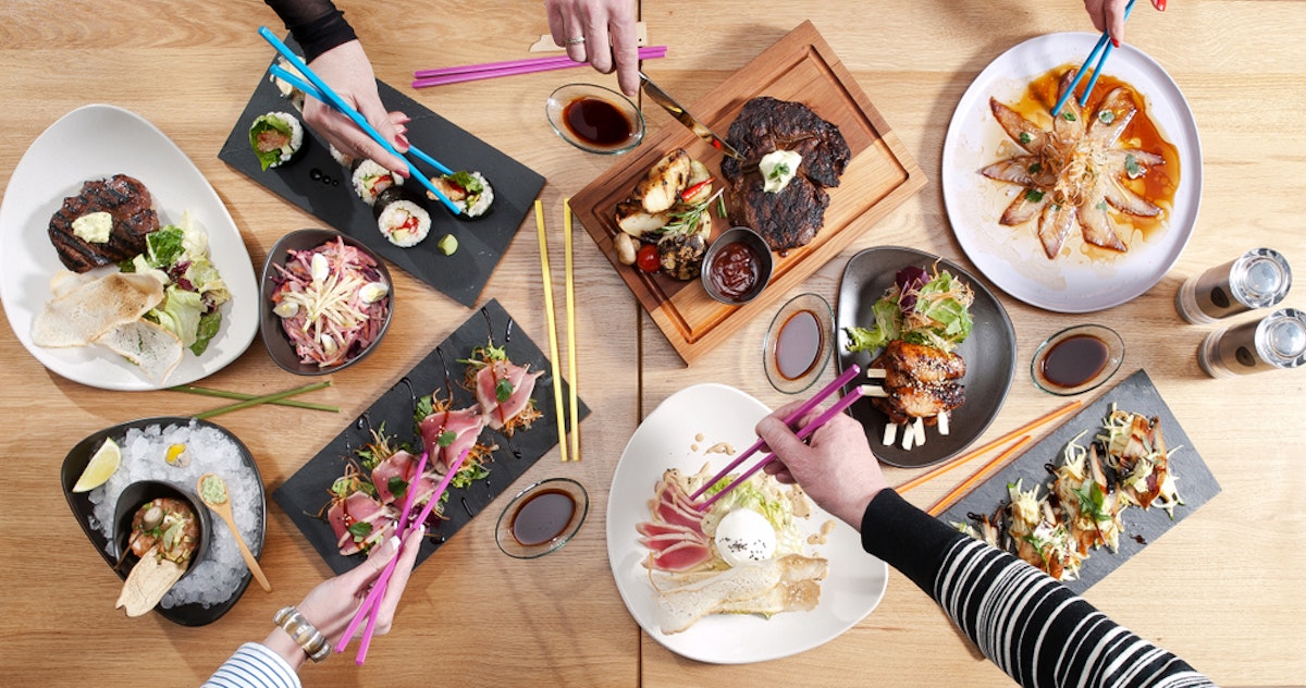 Aerial view of a dining table with various dishes, including sushi, grilled meats, salads, and desserts. Multiple hands using chopsticks reach for the food, creating a lively and colorful scene.