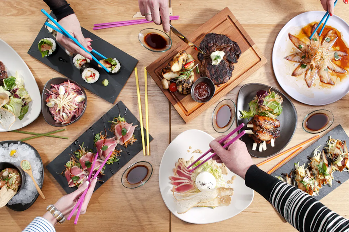 Japanese Food Aerial view of a dining table with various dishes, including sushi, grilled meats, salads, and desserts. Multiple hands using chopsticks reach for the food, creating a lively and colorful scene.
