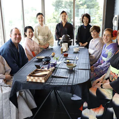 Dining in Kimono A group of nine people in traditional Japanese clothing sit and stand around a table set for a tea ceremony in a bright room with large windows. Everyone is smiling at the camera.