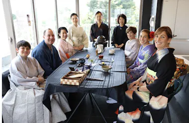 A group of nine people in traditional Japanese clothing sit and stand around a table set for a tea ceremony in a bright room with large windows. Everyone is smiling at the camera.