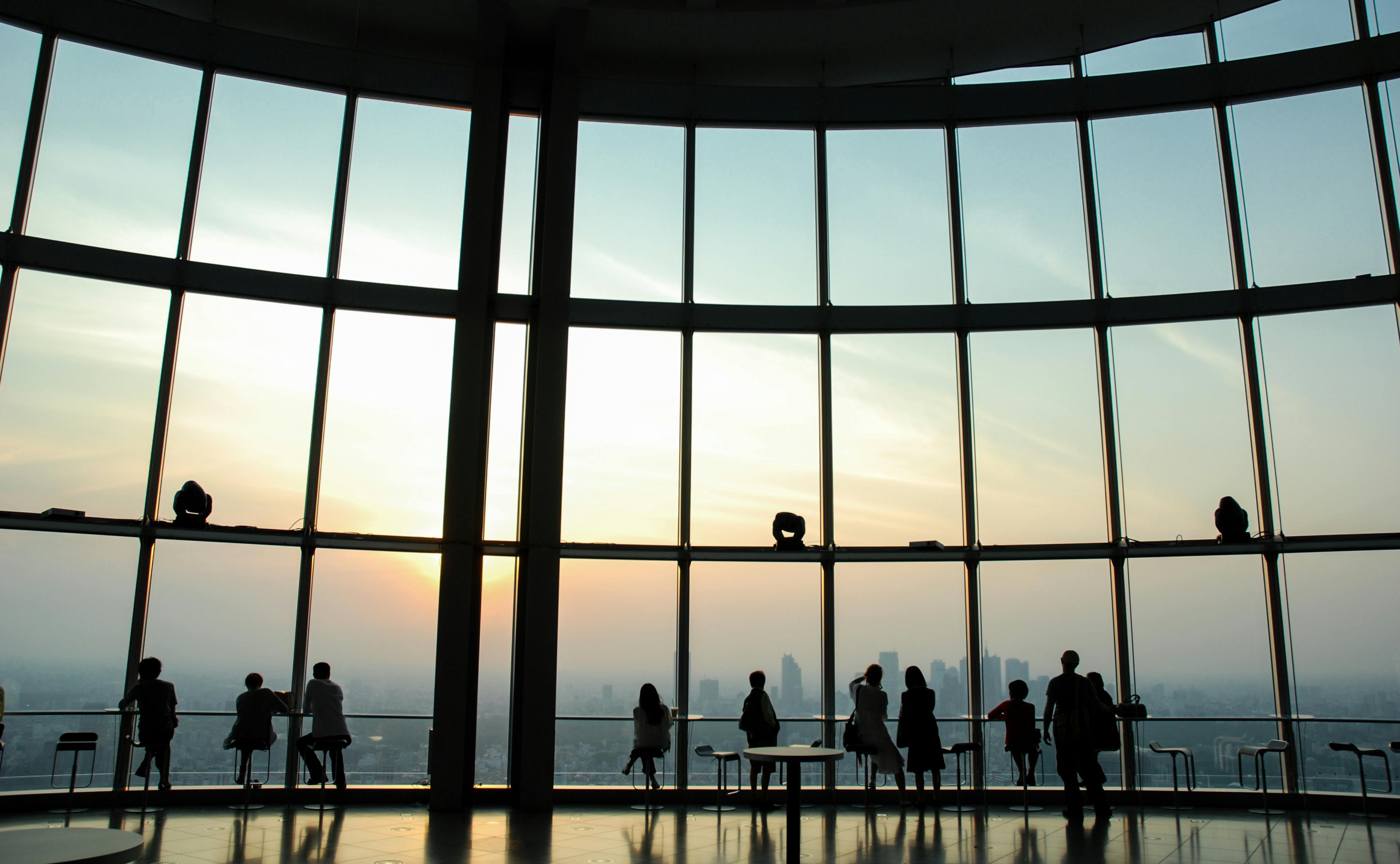 Silhouettes of people standing and sitting by large floor-to-ceiling windows, overlooking a city skyline at sunset. The sky is softly lit, and the scene is peaceful and spacious.